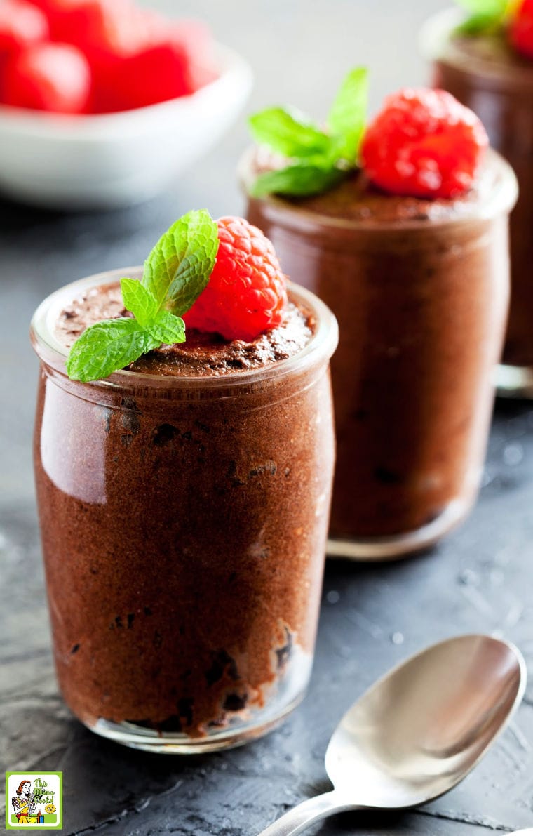 Glasses of chocolate mousse garnished with raspberries and mint with a spoon and bowl of raspberries.