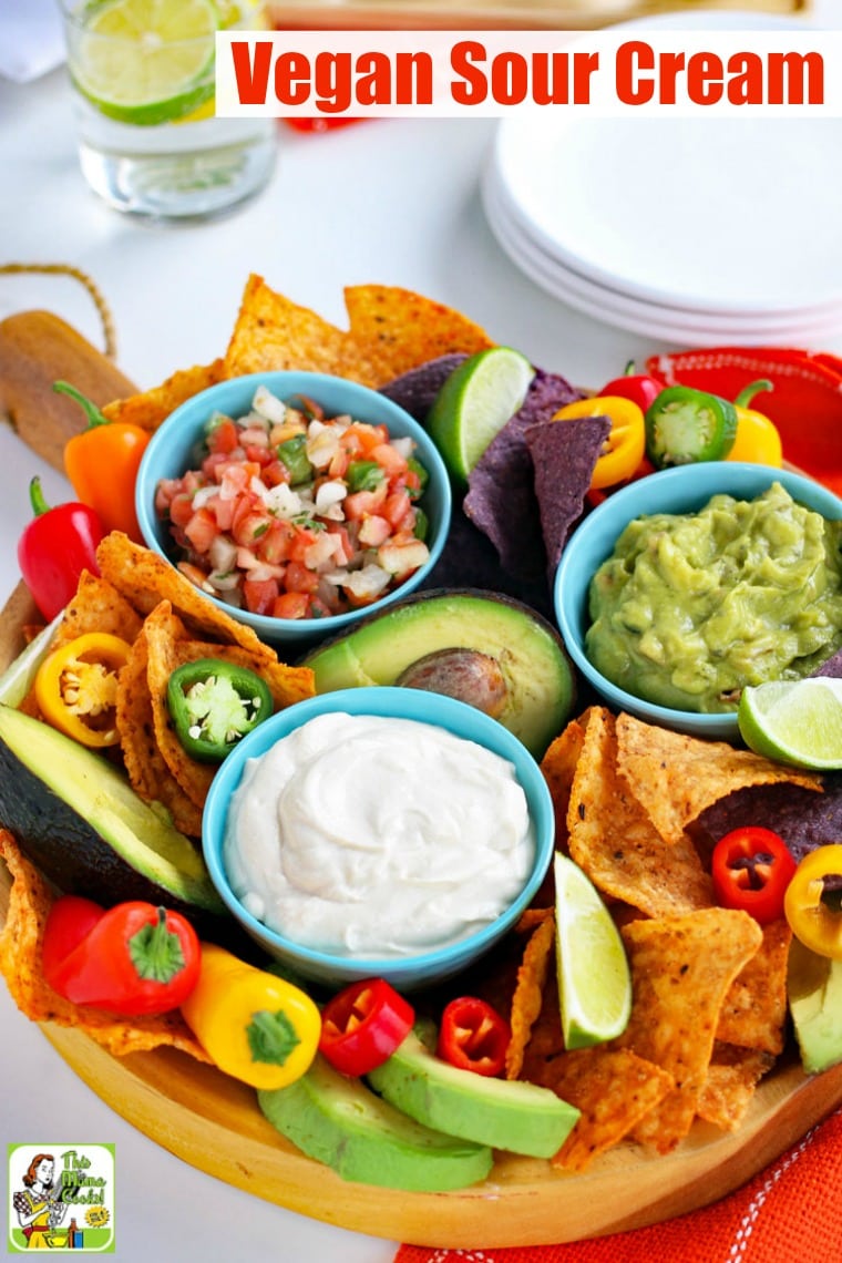 A platter with a small blue bowl of Vegan Sour Cream with slices of limes, tortilla chips, mini sweet peppers, avocados, blue bowls of salsa and guacamole, and white plates.