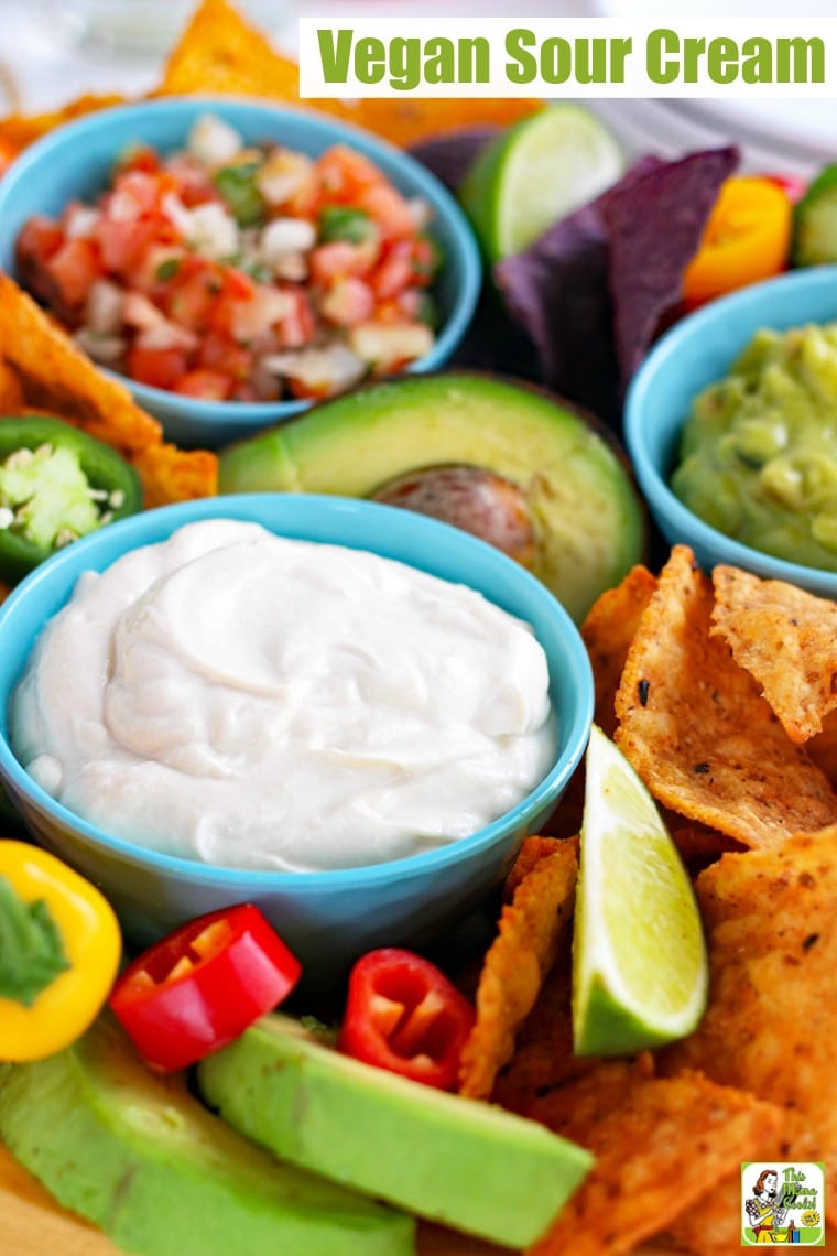 Closeup of blue bowls of creamy vegan sour cream, salsa, and guacamole with slices of lime, tortilla chips, sweet peppers, and avocados.