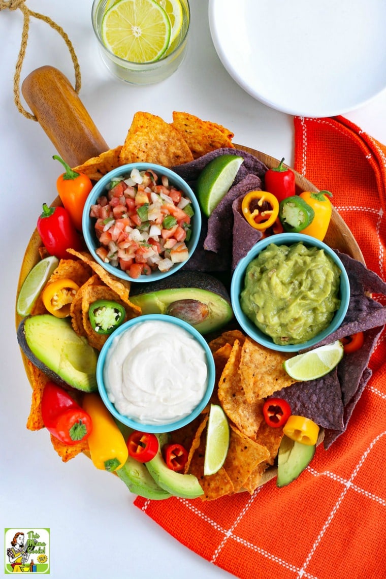 Overhead view of a platter with a bowls of vegan tofu sour cream, salsa, and guacamole with dippers of sweet peppers and tortilla chips along with big bowls of vegan sour cream and a water glass.
