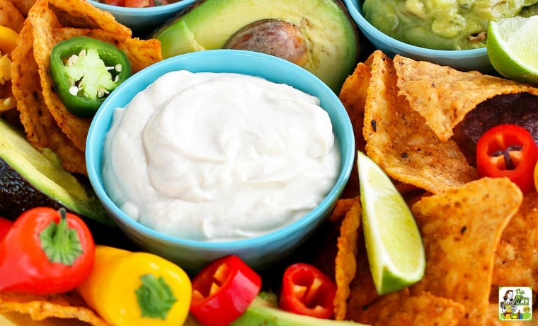 A small blue bowl of Vegan Sour Cream with tortilla chips, mini peppers, avocado, guacamole, and bowls of salsa.