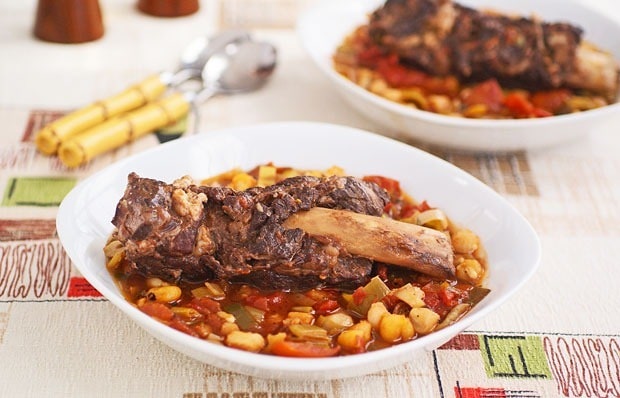 A bowl of braised short ribs on a colorful tablecloth with another bowl and spoons in the background. 