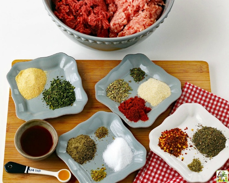 Ingredients for making homemade Italian sasage in small bowls and ceramic trays on a wooden cutting board with a measuring spoon, napkin and a large mixing bowl with ground meat.
