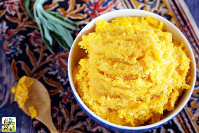 A bowl of Crock-Pot Vegan Mashed Sweet Potatoes with Parsnips, a wooden spoon, and a sprig of sage on a brightly colored tablecloth.