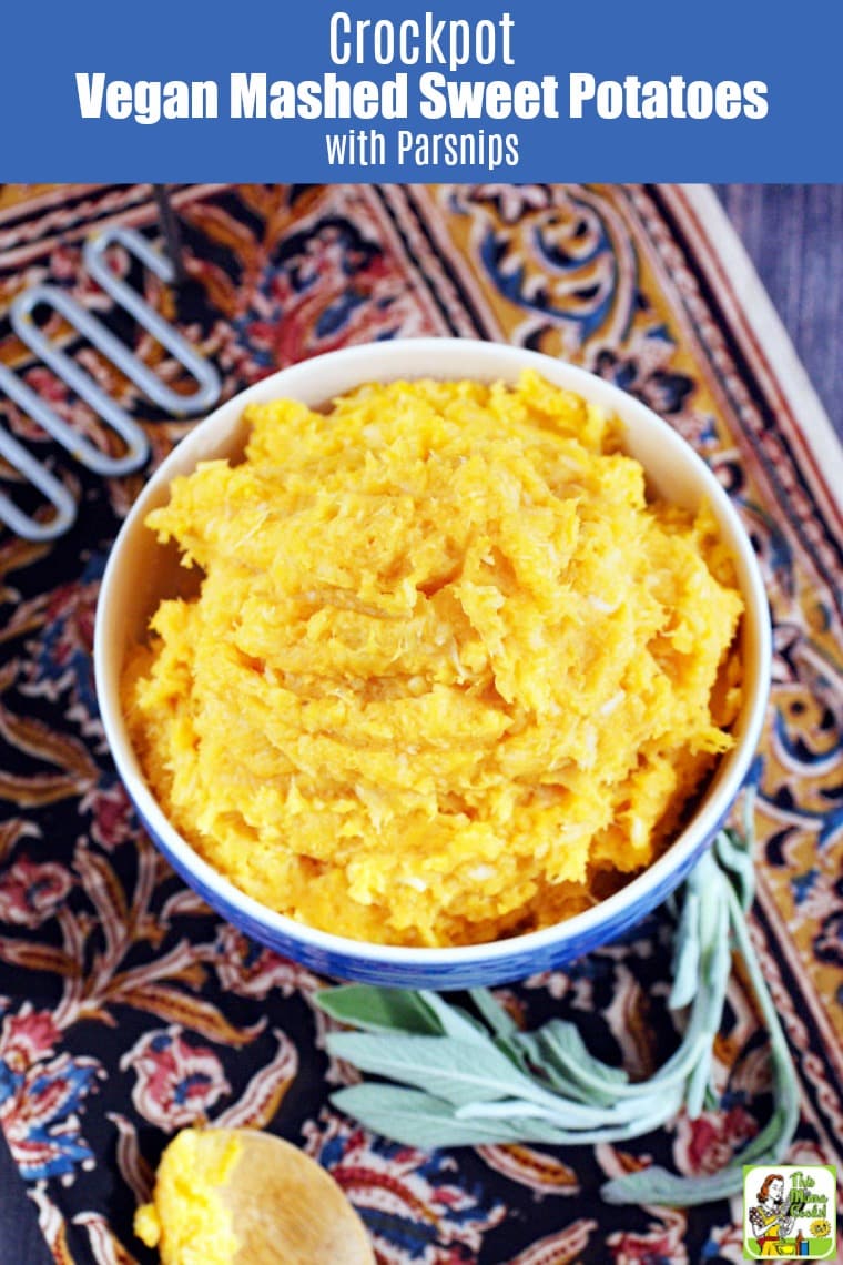 A bowl of Mashed Sweet Potatoes with potato masher on a patterned tablecloth with wooden spoon and sprig of sage.