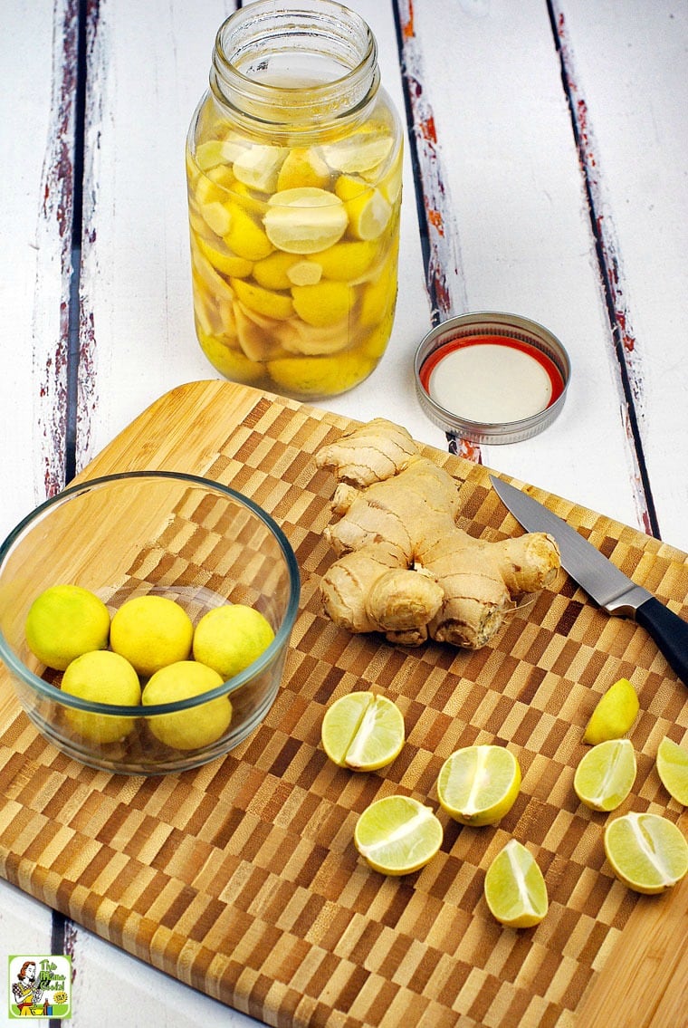 A wooden cutting board with a bowl of limes, ginger and a pairing knife and a jar of infused vodka.