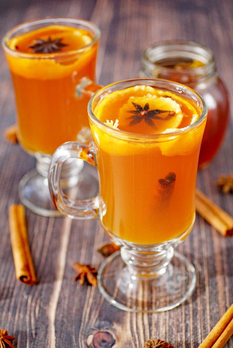Glass mugs of hot tea and whiskey with honey, orange peel, star anise, cloves, cinnamon stick, and a glass jar of honey.