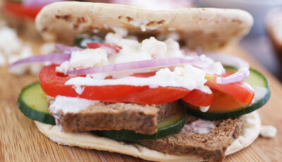 A homemade gyros sandwich with feta, sliced onion, slice cucumber, and tzatziki sauce on a wooden platter.