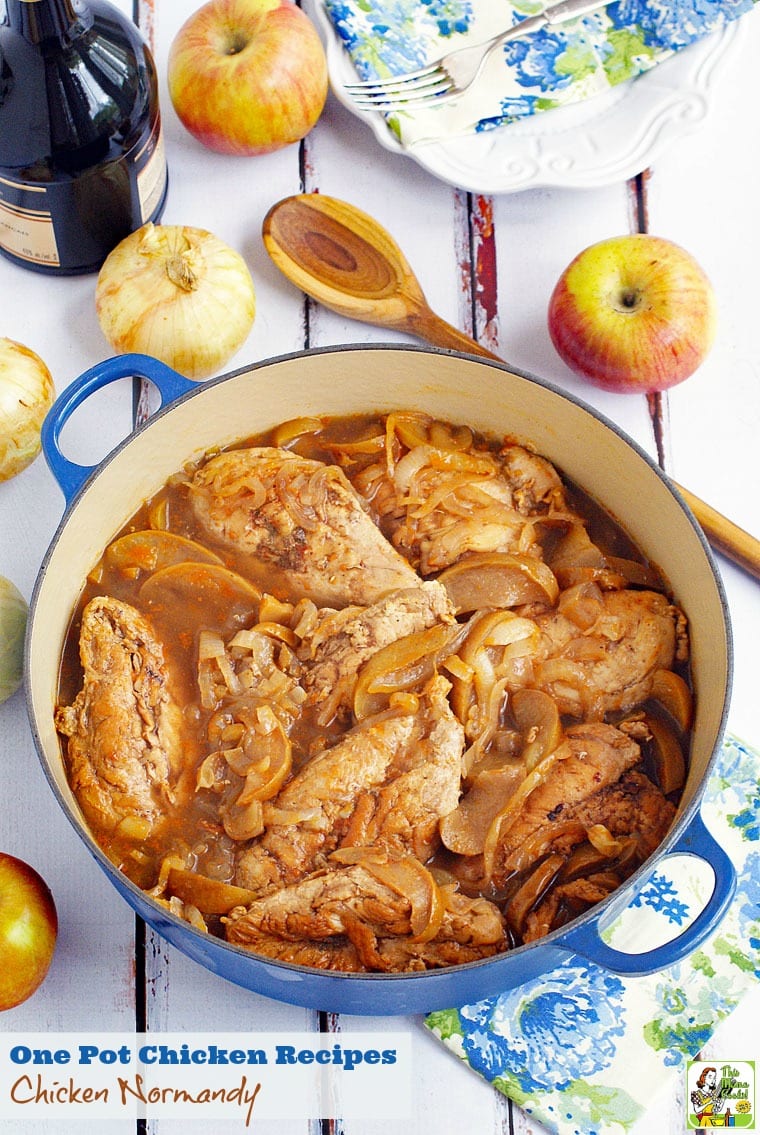 A pot of chicken Normandy made with apples and onions. With apples, onions, a bottle of brandy, floral napkins and a wooden serving spoon. 