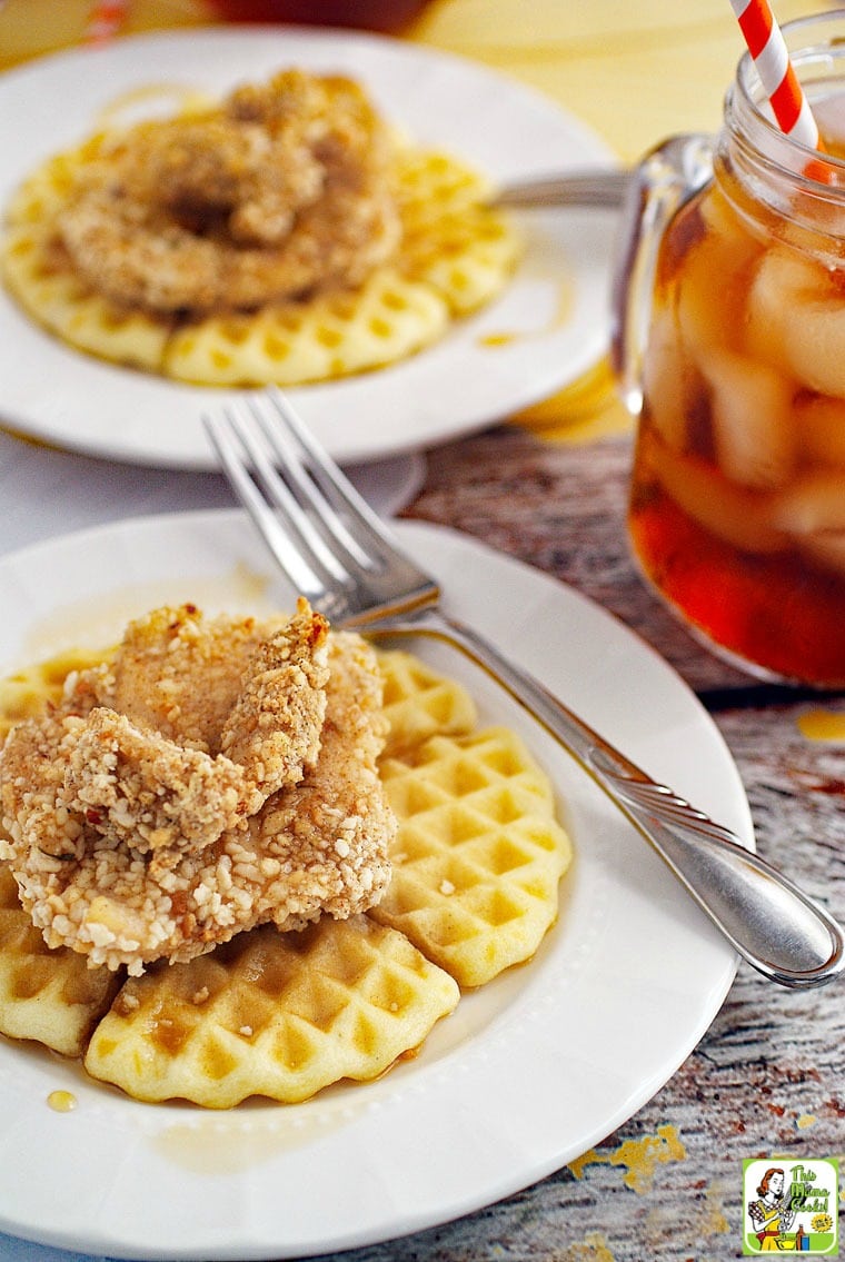 Closeup of chicken and waffles covered in maple syrup on a white plate with a silver fork with a glass of ice tea in the background.