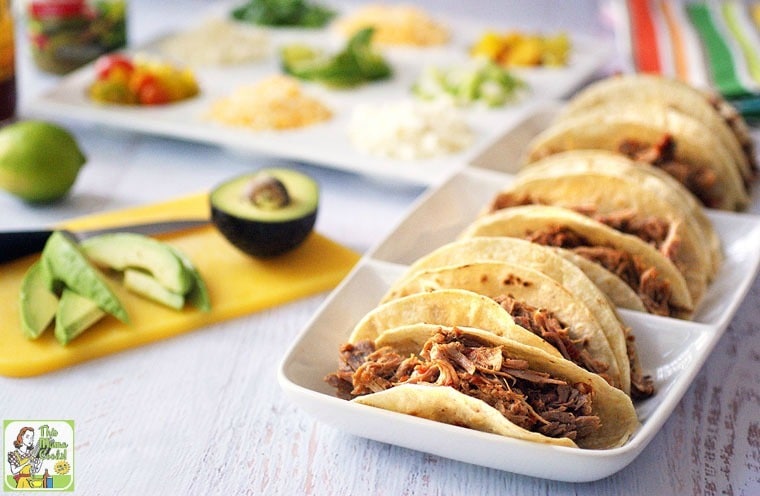 A tray of pulled pork tacos next to a yellow cutting board with avocados.