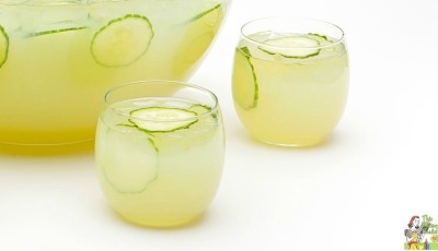 Two glasses and a punch bowl of sparkling green sangria filled with slices of cucumber and grapes.