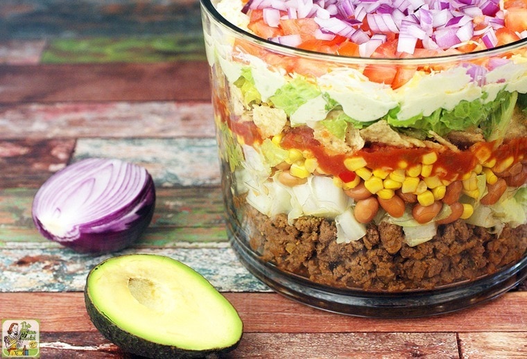 A trifle bowl of layered taco salad with a sliced onion and avocado.