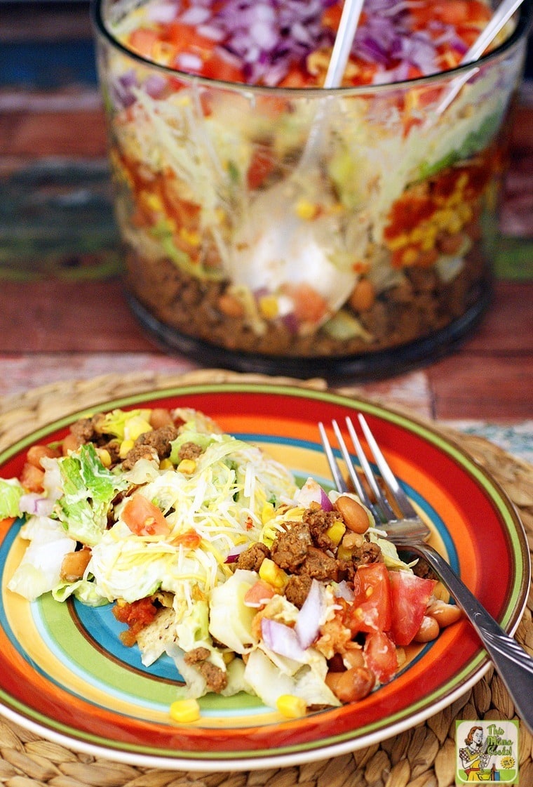 A plate of taco salad on a striped plate with a fork and taco salad in a trifle bowl in the background.