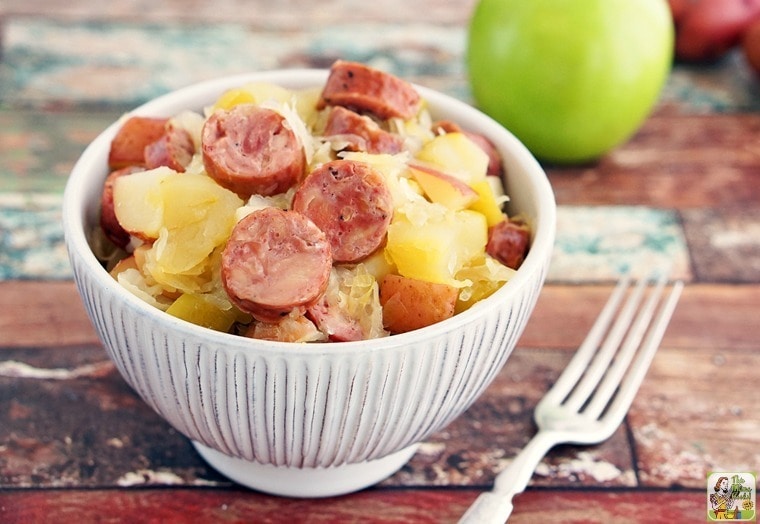 Slow Cooker Sauerkraut and Sausage with Apples and Potatoes in white bowl, with fork and green apples and red potatoes.
