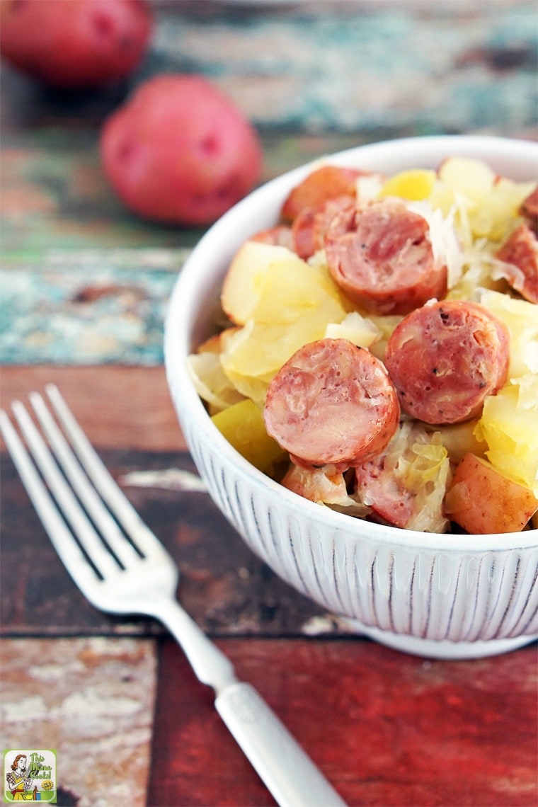 A closeup of slow cooker sauerkraut and sausage with apples and potatoes in a white bowl with fork and red potatoes.