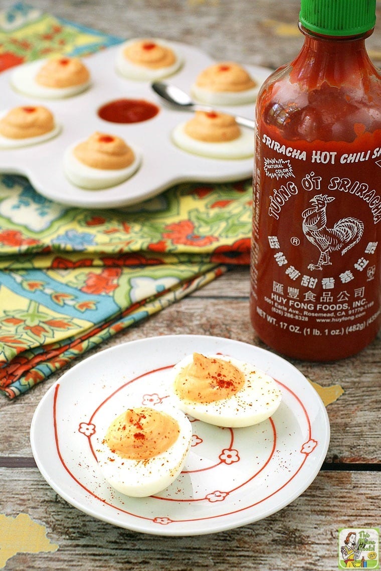 Two deviled eggs on a white plate with a bottle of sriracha sauce and colorful napkins in the background.