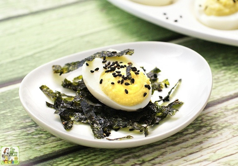 A wasabi deviled egg on a white plate with strips of dried seaweed and black sesame seeds.