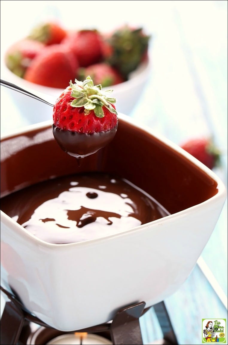 A strawberry on a fondue skewer dipping into a cup of chocolate fondue with a bowl of strawberries in the background.