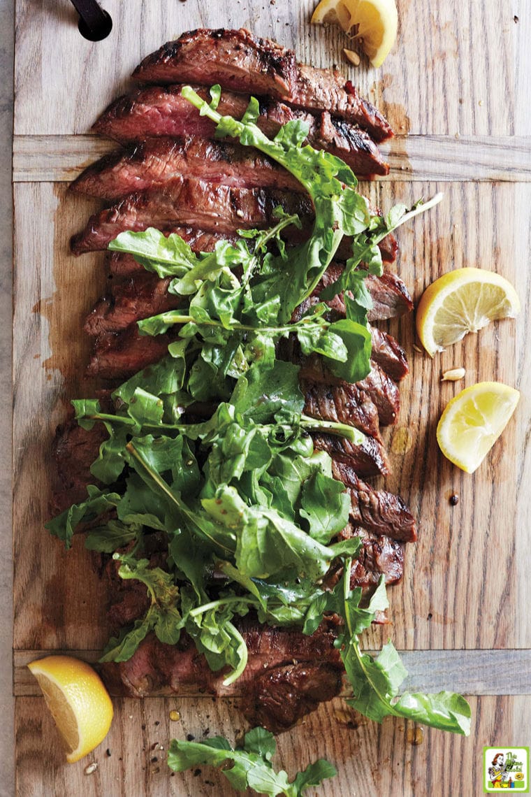 Sliced grillied flank steak with arugula on a wooden cutting board with lemon slices.