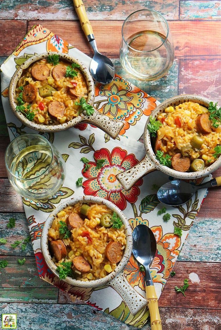 Overhead shot of three bowls of Andouille Sausage Jambalaya on a colorful floral napkin. with a glass of white white and soup spoons.