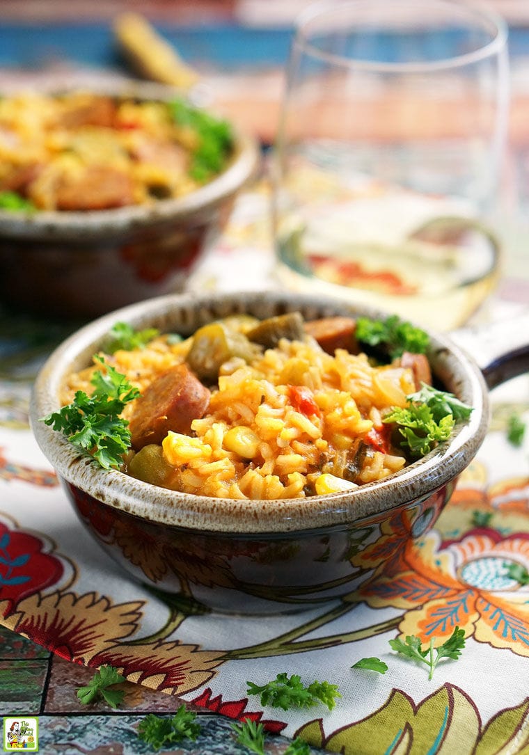 Closeup of a brown lug bowl of Andouille sausage jambalaya on a floral napkin with another bowl and a wine glass in the background.