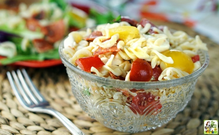 A glass bowl of Caprese Pasta Salad on a woven mat with a plate of salad in the background.
