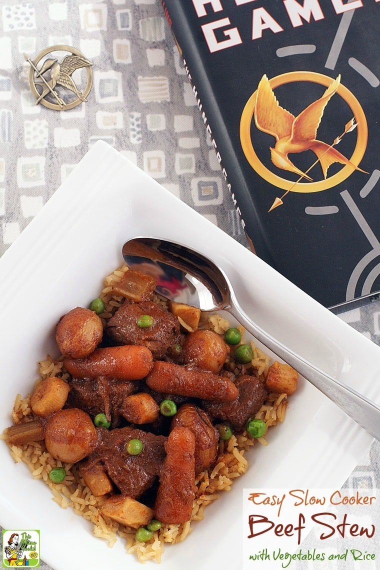 A bowl of slow cooker beef stew with vegetables and rice and The Hunger Games book and pin.