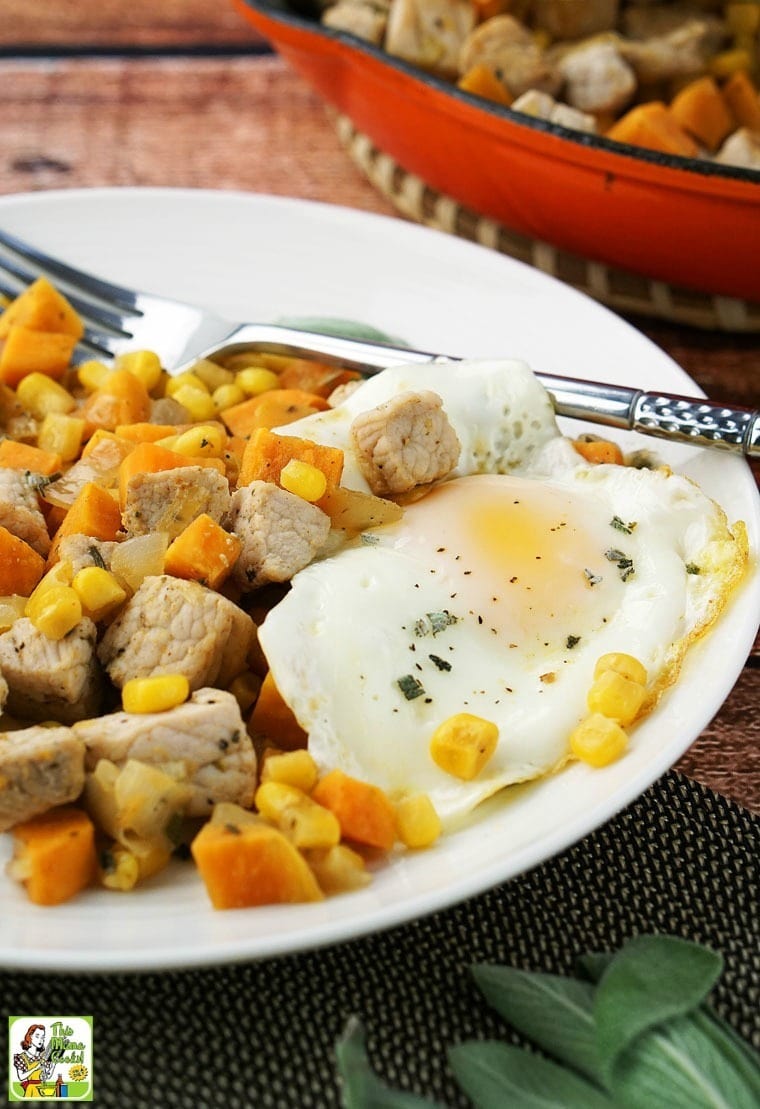 A plate of Sweet Potato Pork Hash with sunny side up eggs, and a fork with a skillet of pork hash in the background