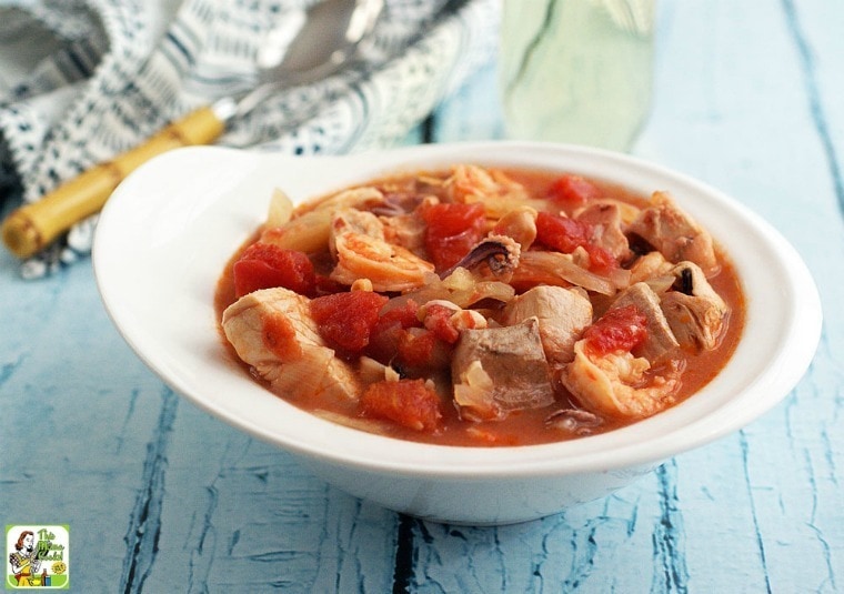 A white bowl of Cioppino Seafood Stew with spoon, napkin, and glass of white wine.