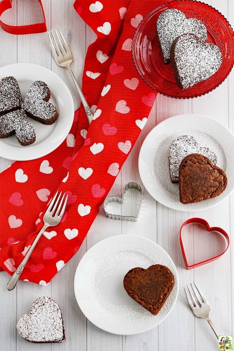 Red and white plates of gluten free fudgy brownies sprinkled with powdered sugar. The brownies are surrounded by heart shaped cookie cutters, forks, and a red dish towel with a pink and white heart pattern. 