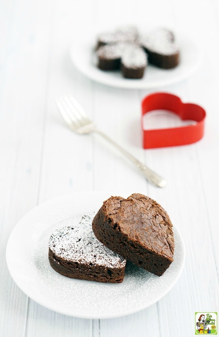 A white plate with heart-shaped gluten-free fudgy brownies. A fork, more brownies, and a heart shaped cookie cutter in the background. 