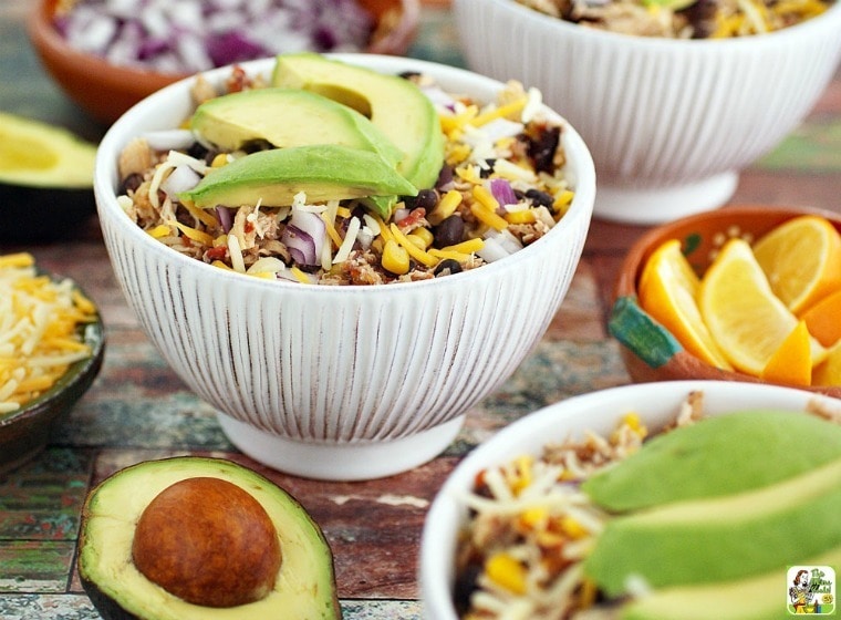Bowls of Slow Cooker Mexican Shredded Chicken with toppings of avocado, cheese, and onions and bowls of lemons.