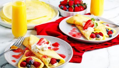 Plates of rice flour crepes with raspberries, blueberries, whipped cream, and berry drizzle with red napkins and glasses of orange juice.