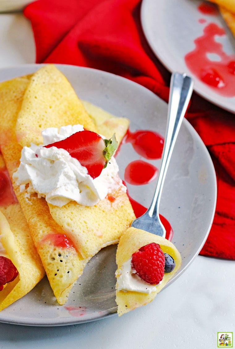 A forkful of piece of crepe with raspberry on a plate of crepes, berries, and whipped creme.