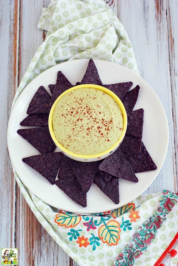 Easy Edamame Hummus in a bowl served with blue tortilla chips on a white plate with a decorative floral tea towel in the background.