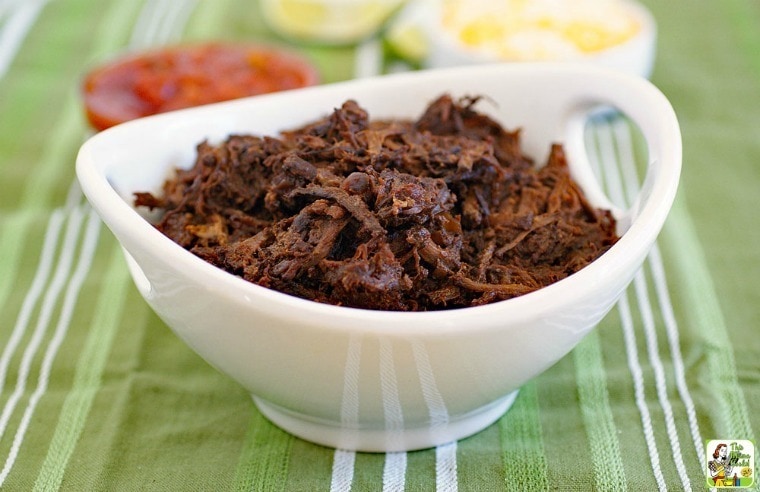 A white bowl of slow cooker goat on a green tablecloth with condiments in the background.