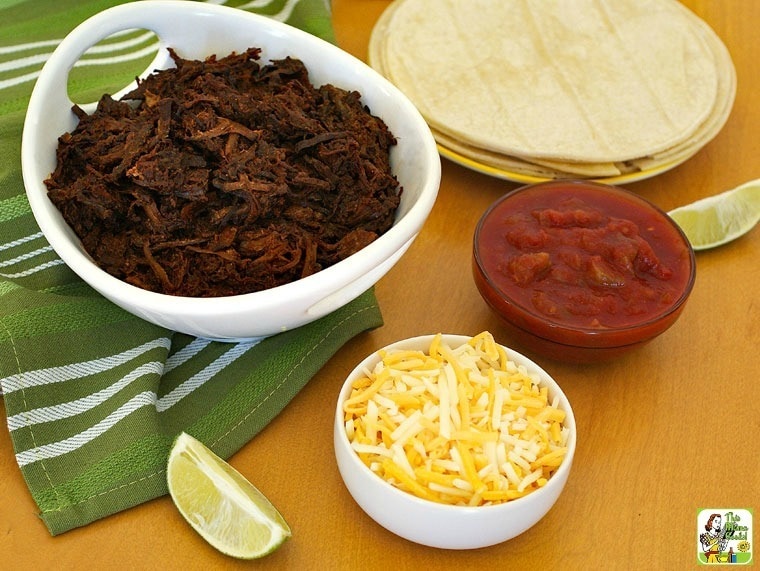 A bowl of shredded slow cooker goat with tortillas, bowls of salsa and cheese, and slices of lime.
