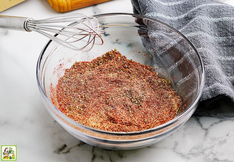 A glass bowl of homemade taco seasoning mix with a wire whisk and gray dish towel.