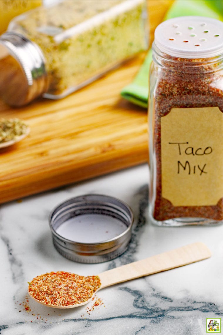 Bottle of taco seasoning mix, a spoon of taco mix, and a wooden cutting board on a marble counter top.
