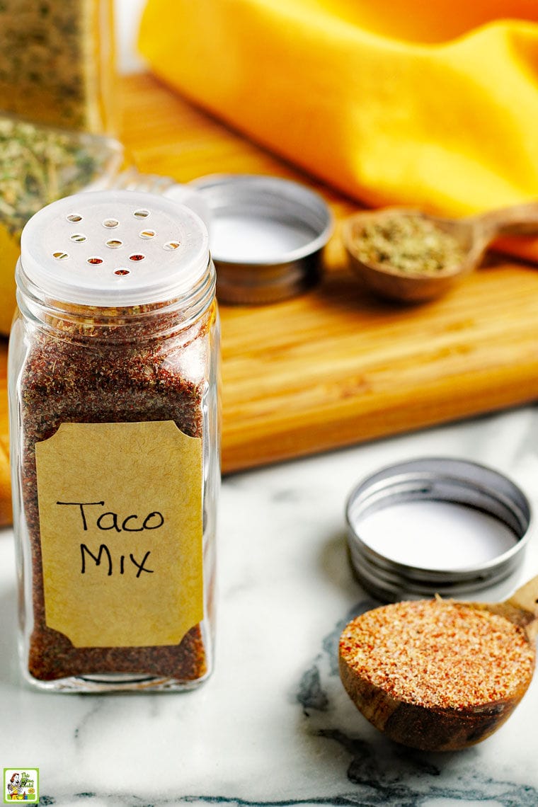 Bottle of taco mix, a wooden spoon of taco mix, and a wooden cutting board on a marble counter top with a yellow napkin.