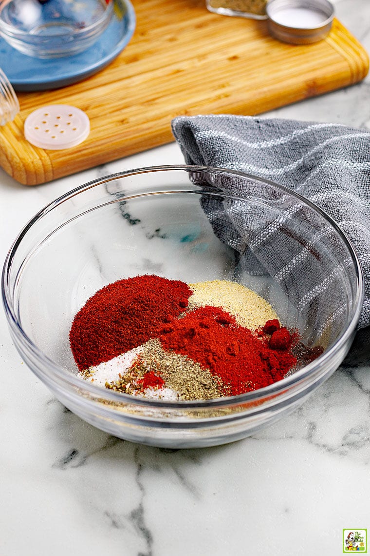 Making homemade taco seasoning in a glass bowl with a variety of seasonings with a gray dish towel and a wooden cutting board.
