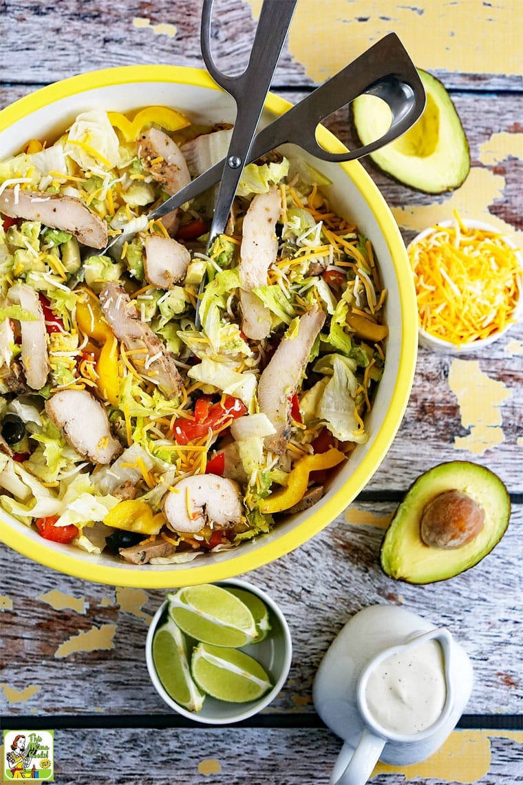 A large yellow bowl of Chicken Fajita Salad with cheese, avocado, limes, and a pitcher of salad dressing. 