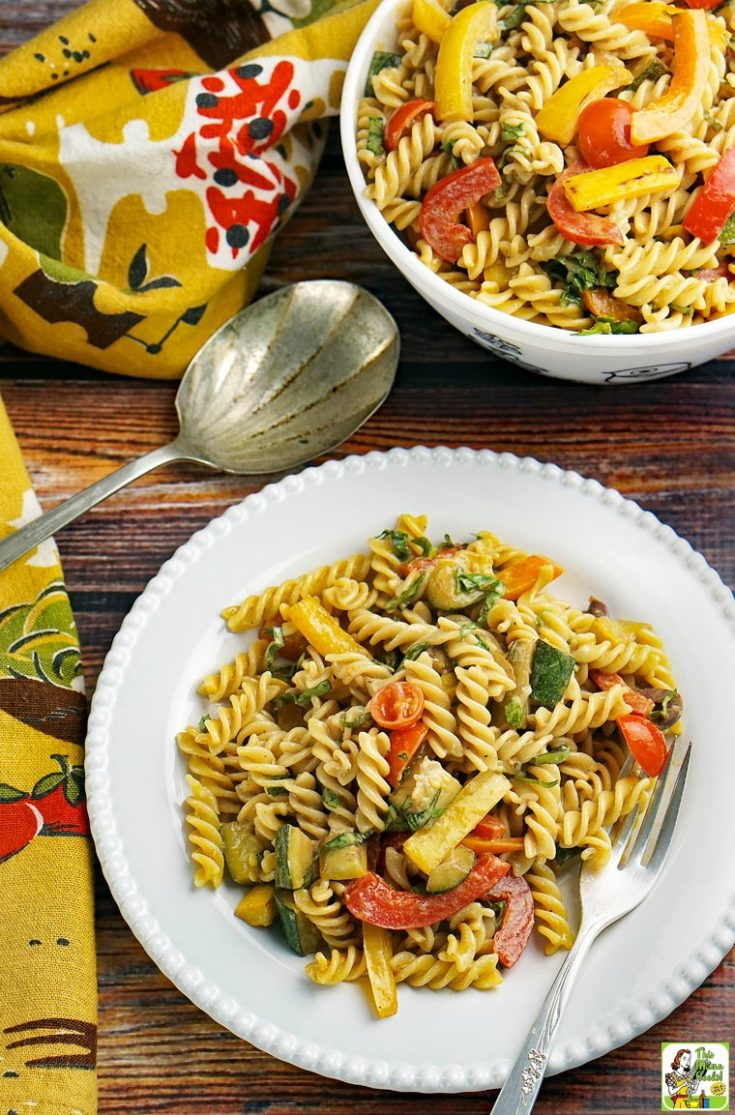 A white plate of pasta salad with roasted vegetables with a bowl of pasta salad, a serving spoon, and napkins.