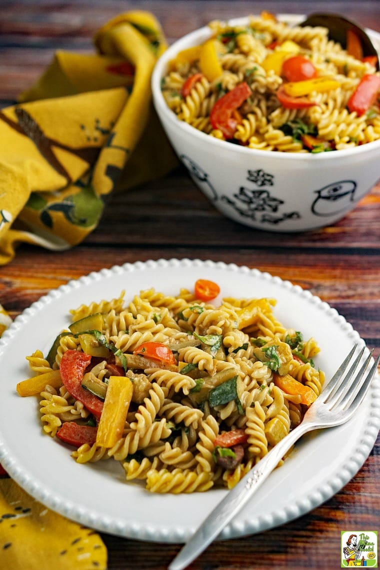 A white plate of pasta salad with roasted vegetables with a bowl of pasta salad in the background. 
