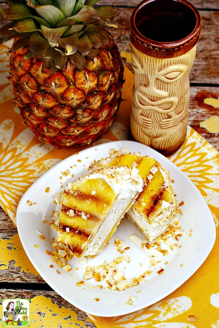 Grilled pineapple tropical ice cream sandwich on a white plate with toasted coconut with a whole pineapple and a tiki glass.