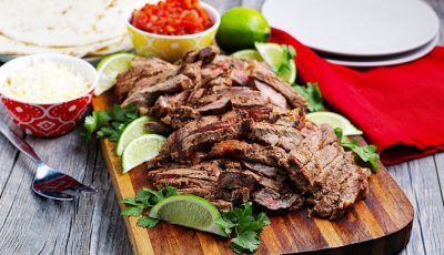 Sliced carne asada meat with limes and cilantro on a wooden cutting board with taco fixings in the background.