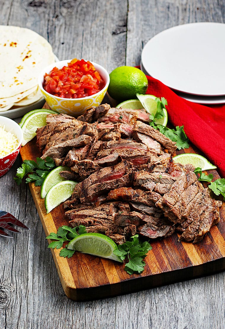 Sliced grilled carne asada meat on a wooden board with a bowl of salsa and slices of lime, white plates, a red napkin, and a pile of flour tortillas.