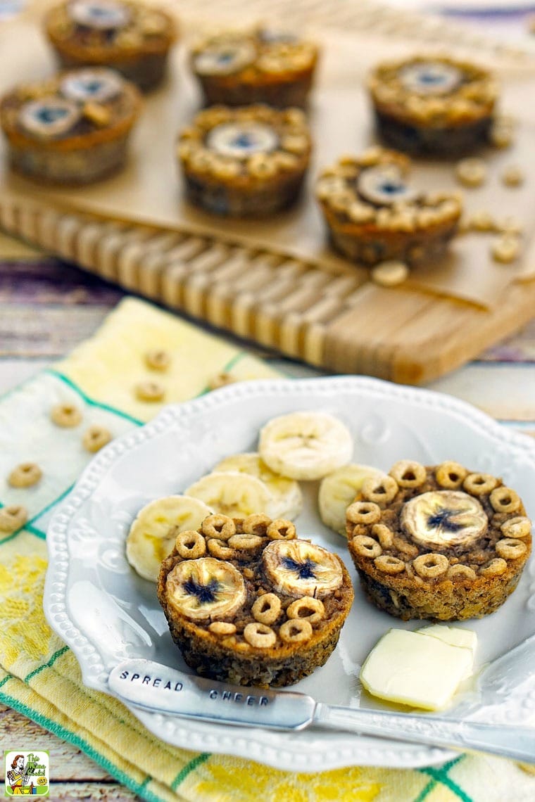 Gluten free banana muffins with Cheerios, banana slices, pats of butter, and a knife on a dish towel.