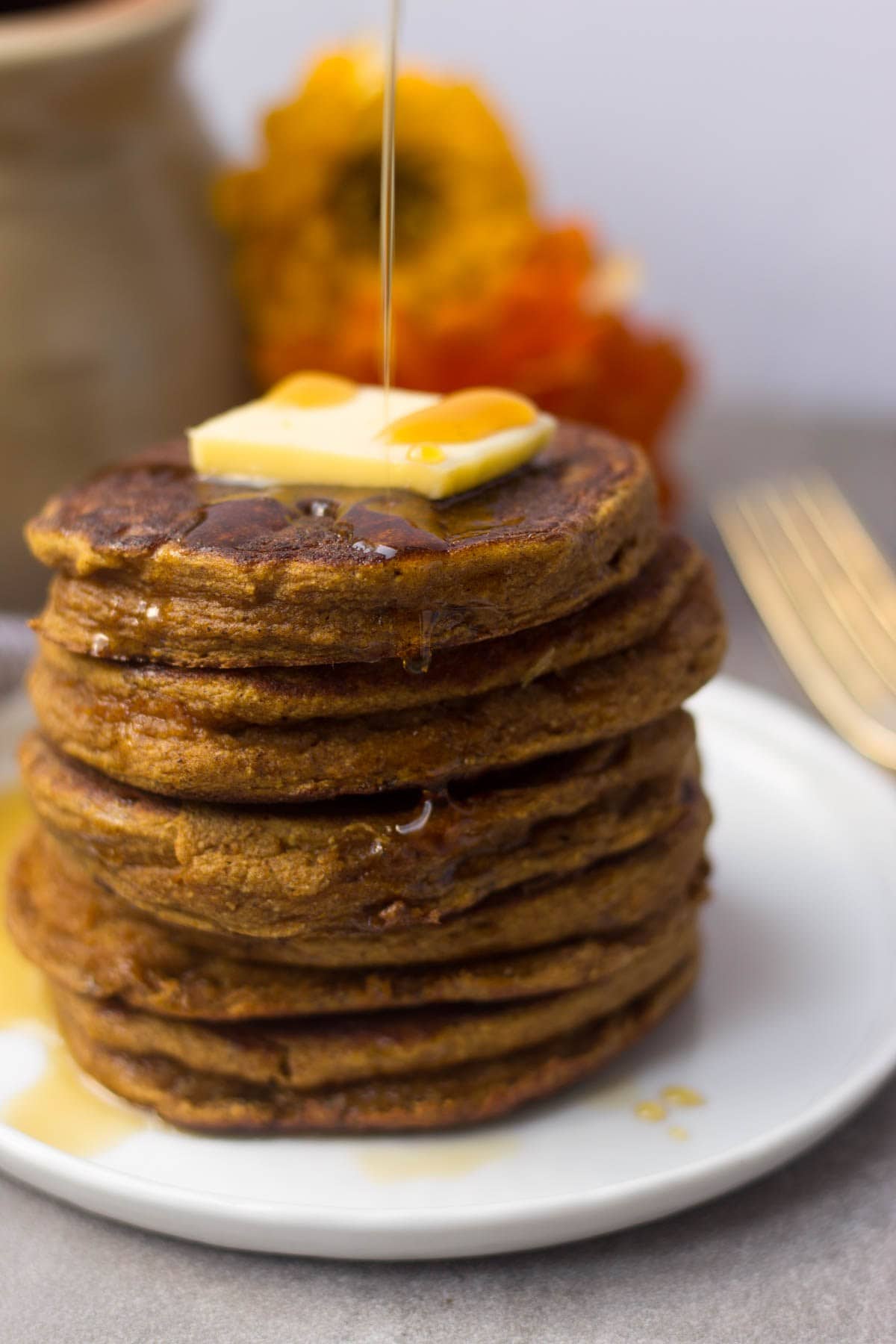 A tall stack of Paleo Pumpkin Protein Pancakes with a pour of syrup.